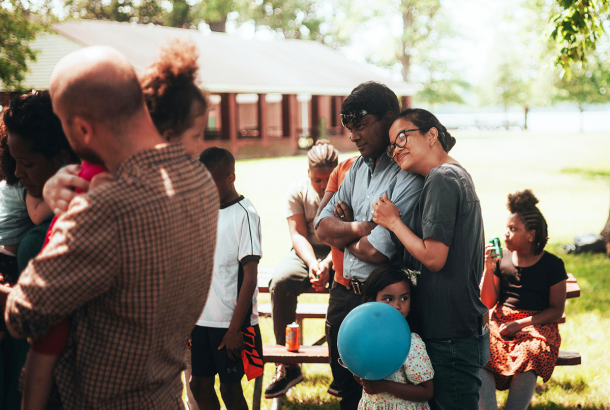 Women with her head on her husbands shoulder smiling with her daughter next to her holding a balloon. Part of a diverse celebration with people praying.